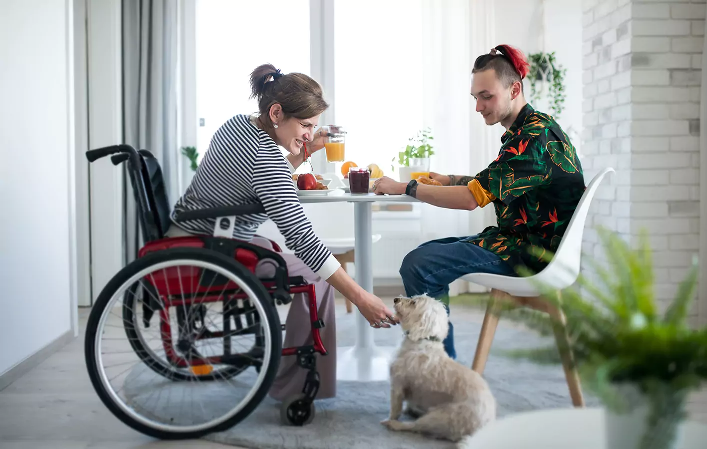 A disabled woman sitting in her wheelchair while reaching for the puppy with her support worker in the dining area